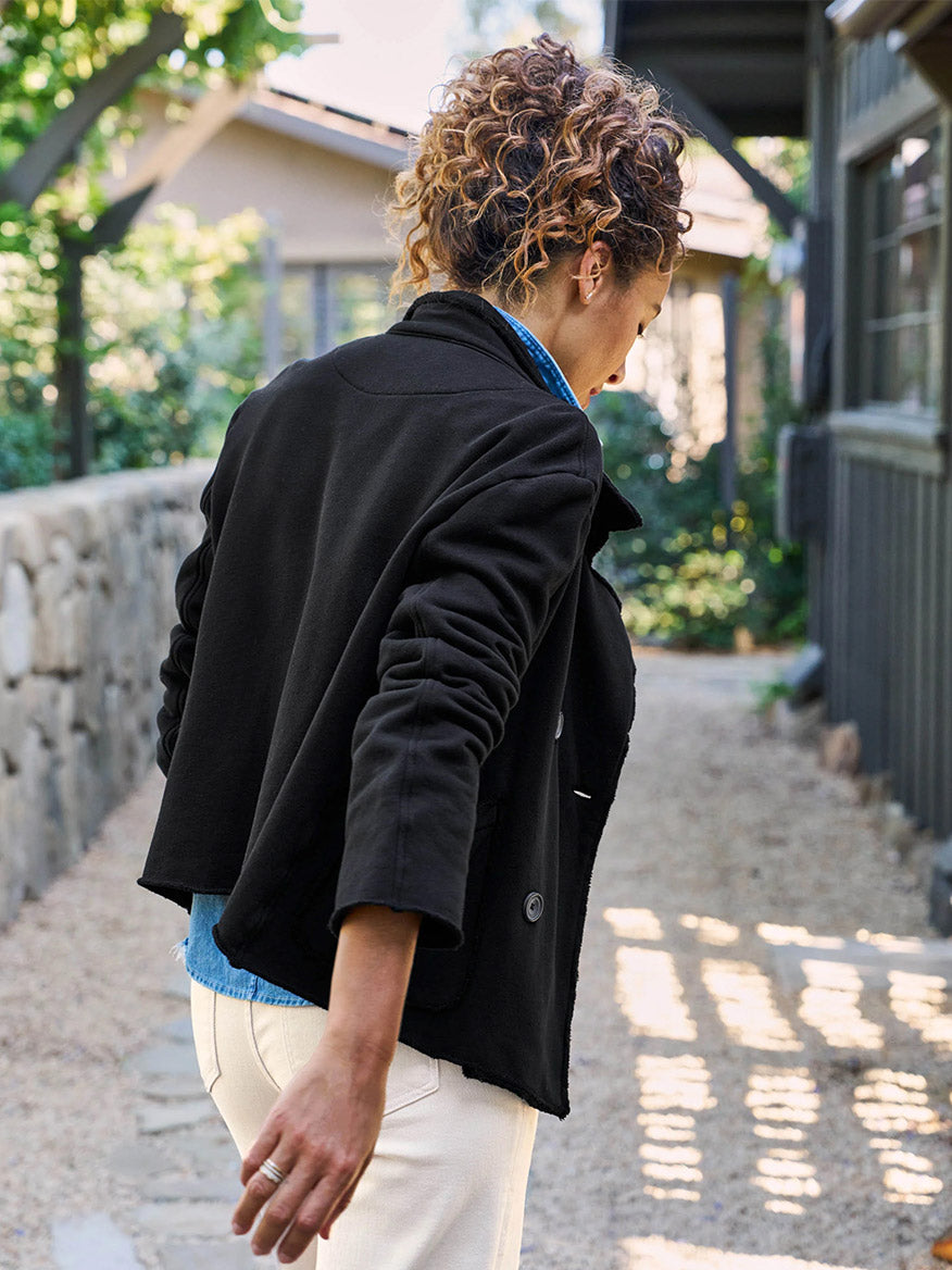 A person with curly hair in a Frank & Eileen Mini Belfast Crop Peacoat in Black Fleece strolls outdoors on a stone path beside a building, basking in the sunlight filtering through the trees. Their attire adds an element of relaxed style to this scenic outing.