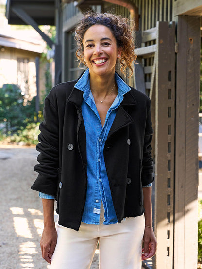 A person with curly hair smiles outdoors, wearing a black Frank & Eileen Mini Belfast Crop Peacoat in fleece over a blue shirt and white pants.