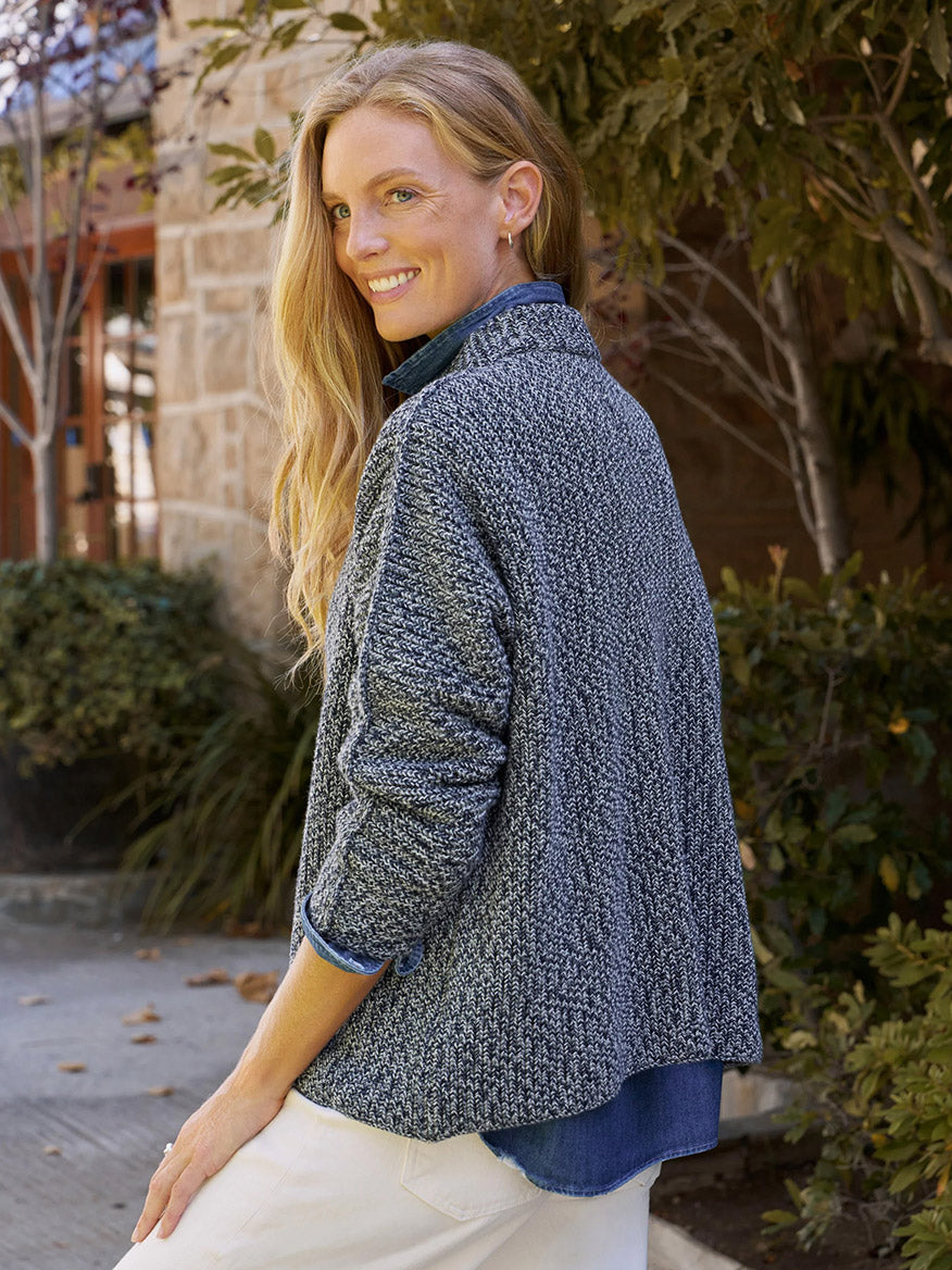 Woman with long blonde hair smiles over her shoulder, wearing the Frank & Eileen Montecito Sweater in Gray and Navy Marl, with greenery and a building behind her.