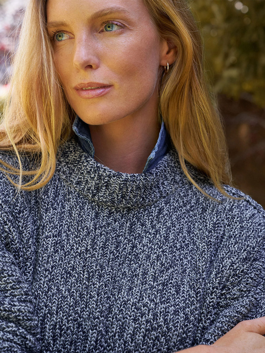 A woman with long blonde hair wears a relaxed-fit Frank & Eileen Montecito Sweater in Gray and Navy Marl over a blue shirt, standing outdoors with greenery behind her.