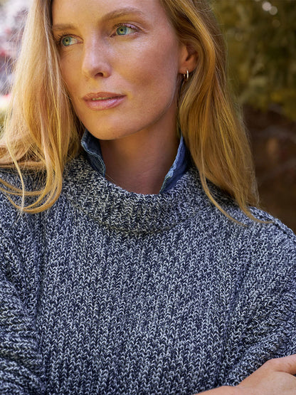 A woman with long blonde hair wears a relaxed-fit Frank & Eileen Montecito Sweater in Gray and Navy Marl over a blue shirt, standing outdoors with greenery behind her.