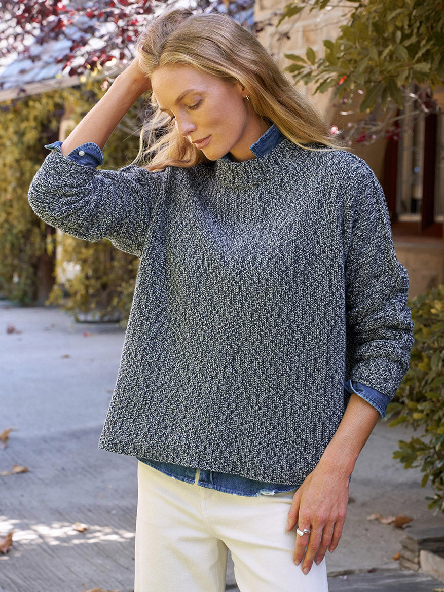 Woman outdoors in a relaxed fit Frank & Eileen Montecito Sweater in Gray and Navy Marl over a blue shirt and white pants, touching her hair. Trees and building in the background.