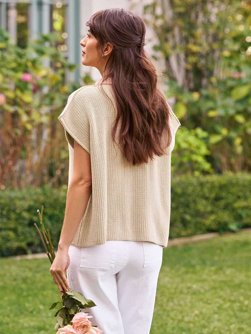 A woman with long brown hair, dressed in the Frank & Eileen Montecito Italian Sweater Vest in Sand and white pants, stands outdoors holding a small bouquet of flowers, facing away from the camera.