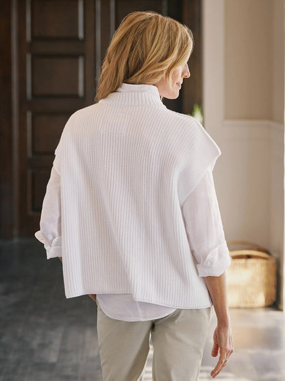 A woman with blonde hair stands indoors facing away, wearing the Frank & Eileen Montecito Italian Sweater Vest in White over a white shirt and beige pants.
