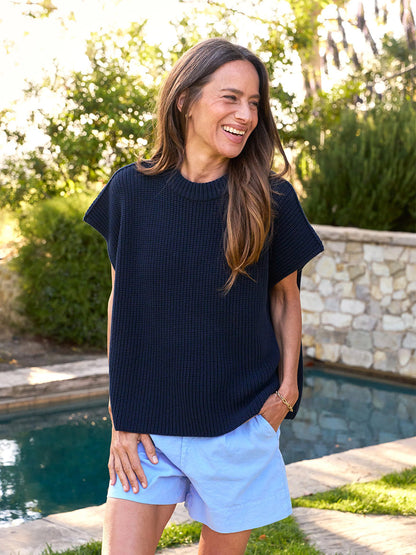 By a pool, a woman smiles in the serene setting, wearing a Frank & Eileen Montecito Italian Sweater Vest in Marine and light blue shorts. Framed by trees and a stone wall, her look captures the charm of classic Pure Italian Cotton style.