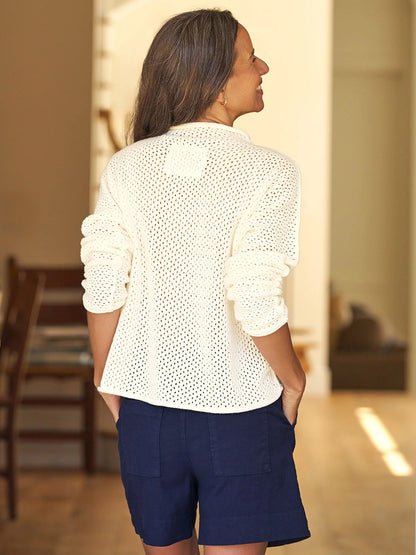 A person with long hair stands indoors facing away, wearing the Frank & Eileen Monterey Pointelle Rolled Funnel Neck Sweater in Ivory and navy shorts, hands tucked casually in their pockets.