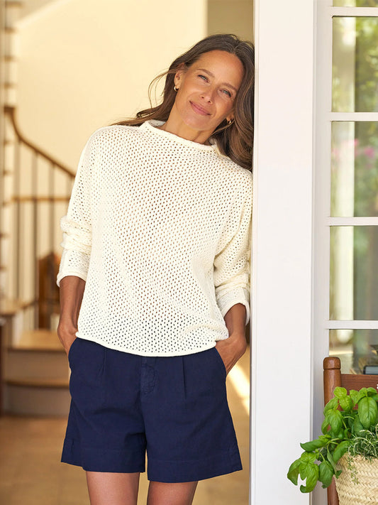 A woman in a Frank & Eileen Monterey Pointelle Rolled Funnel Neck Sweater in Ivory and navy shorts stands indoors, leaning against a door frame and smiling. Spiral stairs and a chair with a plant are visible in the background.