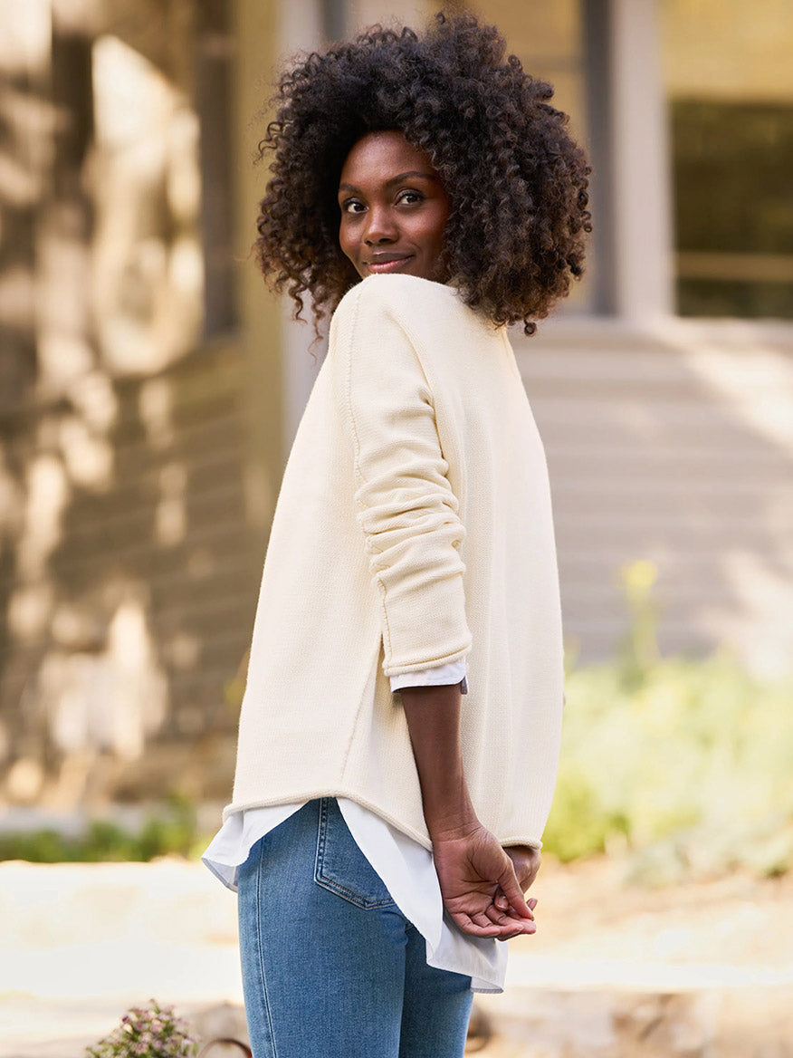 A person with curly hair is standing outdoors, wearing a Frank & Eileen Monterey Rolled Funnel Neck Sweater in cream and blue jeans, looking back over their shoulder in the crisp breeze.