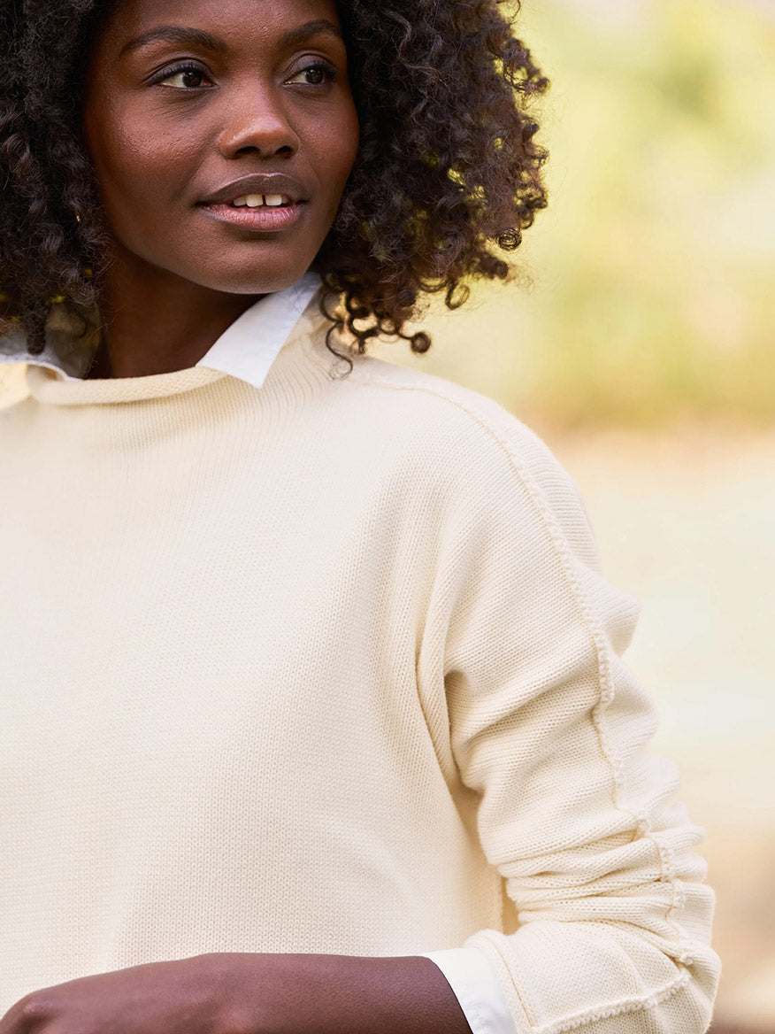 A person with curly hair is wearing a Frank & Eileen Monterey Rolled Funnel Neck Sweater in cream over a white collared shirt, smiling slightly against the blurred backdrop of Monterey outdoors. This machine-washable outfit exudes effortless style and comfort.