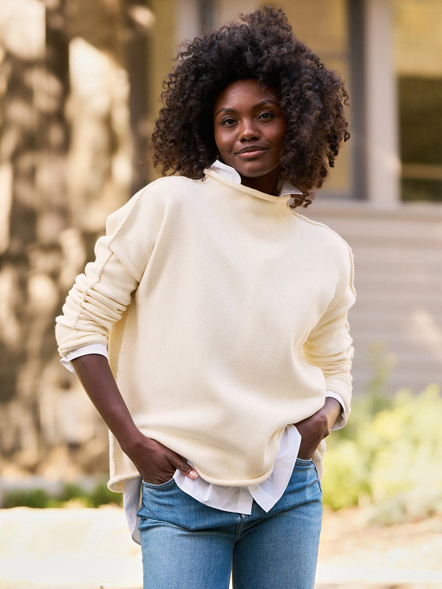 A person stands outside, wearing the Frank & Eileen Monterey Rolled Funnel Neck Sweater in Cream over a white shirt and blue jeans. Their curly hair frames a slight smile, perfectly complementing the relaxed vibe ideal for a Monterey stroll. Plus, the ensemble is machine-washable for easy care.