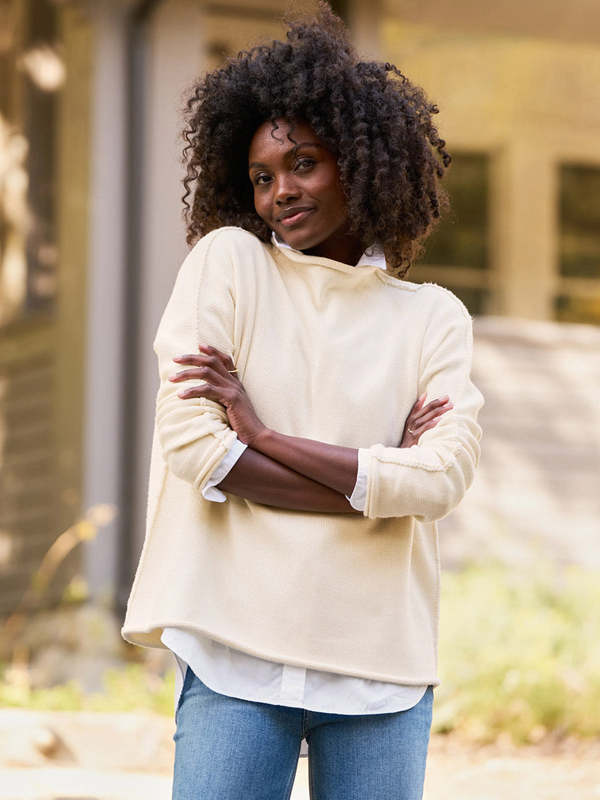 A woman with curly hair stands outdoors in a Monterey landscape, wearing a Frank & Eileen Monterey Rolled Funnel Neck Sweater in Cream over a white shirt and blue jeans. Her arms are crossed, and she has a neutral expression. The sweater is conveniently machine-washable for easy care.