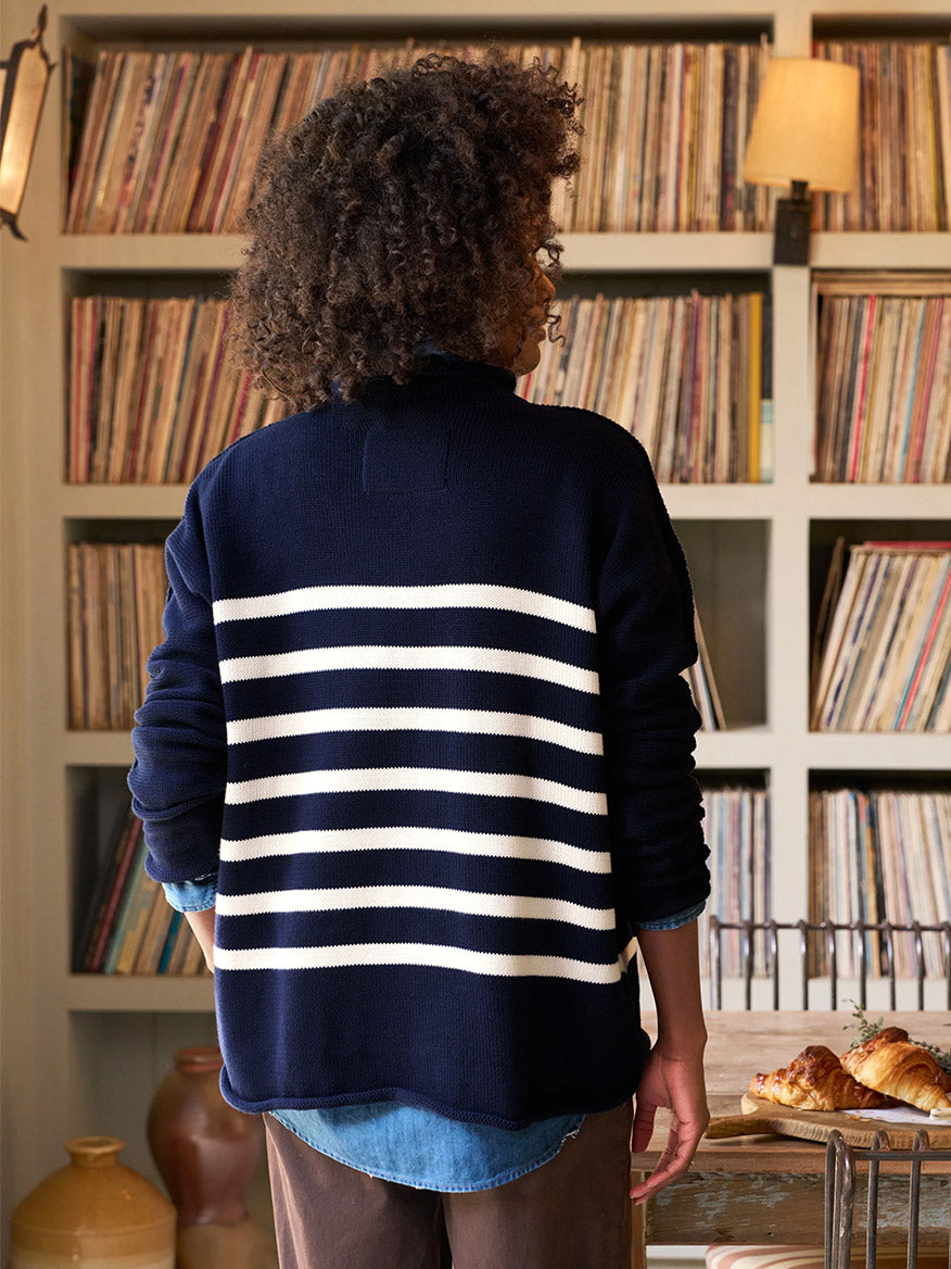 A person wearing a Frank & Eileen Monterey Rolled Funnel Neck Sweater in Navy & Cream Stripe stands in front of a shelf filled with vinyl records, while a nearby table holds an assortment of pastries. The Italian cotton fabric and exposed seam details enhance the cozy setting with a touch of sophistication.