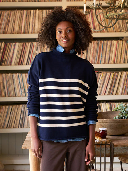 A person with curly hair wears a Frank & Eileen Monterey Rolled Funnel Neck Sweater in Navy & Cream Stripe, standing indoors. Shelves filled with records create a cozy backdrop.