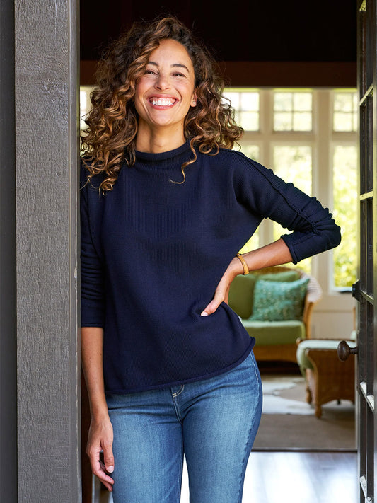 A person with curly hair stands in a doorway wearing a cozy Frank & Eileen Monterey Rolled Funnel Neck Sweater in Navy and jeans, smiling with one hand on their hip. The funnel neck adds style, while a window and green chair in the background complete the warm and inviting scene.