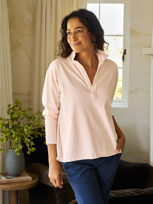A person in a Frank & Eileen Patrick Popover Henley in Petal Heritage Jersey and blue pants stands indoors by a window with one hand in their pocket. A potted plant on a nearby table adds to the cozy scene.