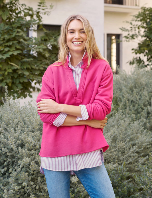 A person smiles, wearing a pink sweater over a Frank & Eileen Patrick Popover Henley in Rose Triple Fleece and raw-hem jeans, standing outside by green shrubs and a building.