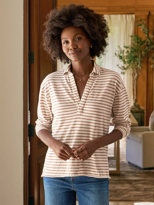 A woman stands indoors, hands together, wearing the Frank & Eileen Patrick Popover Henley in Vintage White & Camel Stripe Heritage Jersey and blue jeans.