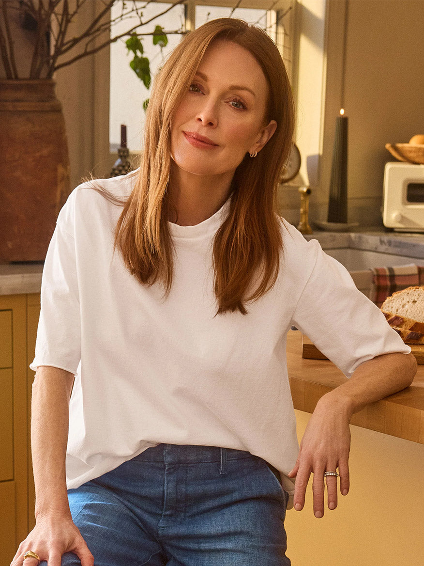 A woman with straight, shoulder-length light brown hair wears the Frank & Eileen Perry NYC Crewneck Tee in White and blue jeans, sitting on a kitchen counter with bread and kitchen items—a hint of New Yorker style at home.