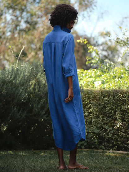 Barefoot on the grass in a garden, a person with curly hair faces away from the camera, effortlessly embodying California chic in the Frank & Eileen Rory Maxi Shirtdress in Cabo Blue, its full-length button-up silhouette and rolled sleeves completing the look.