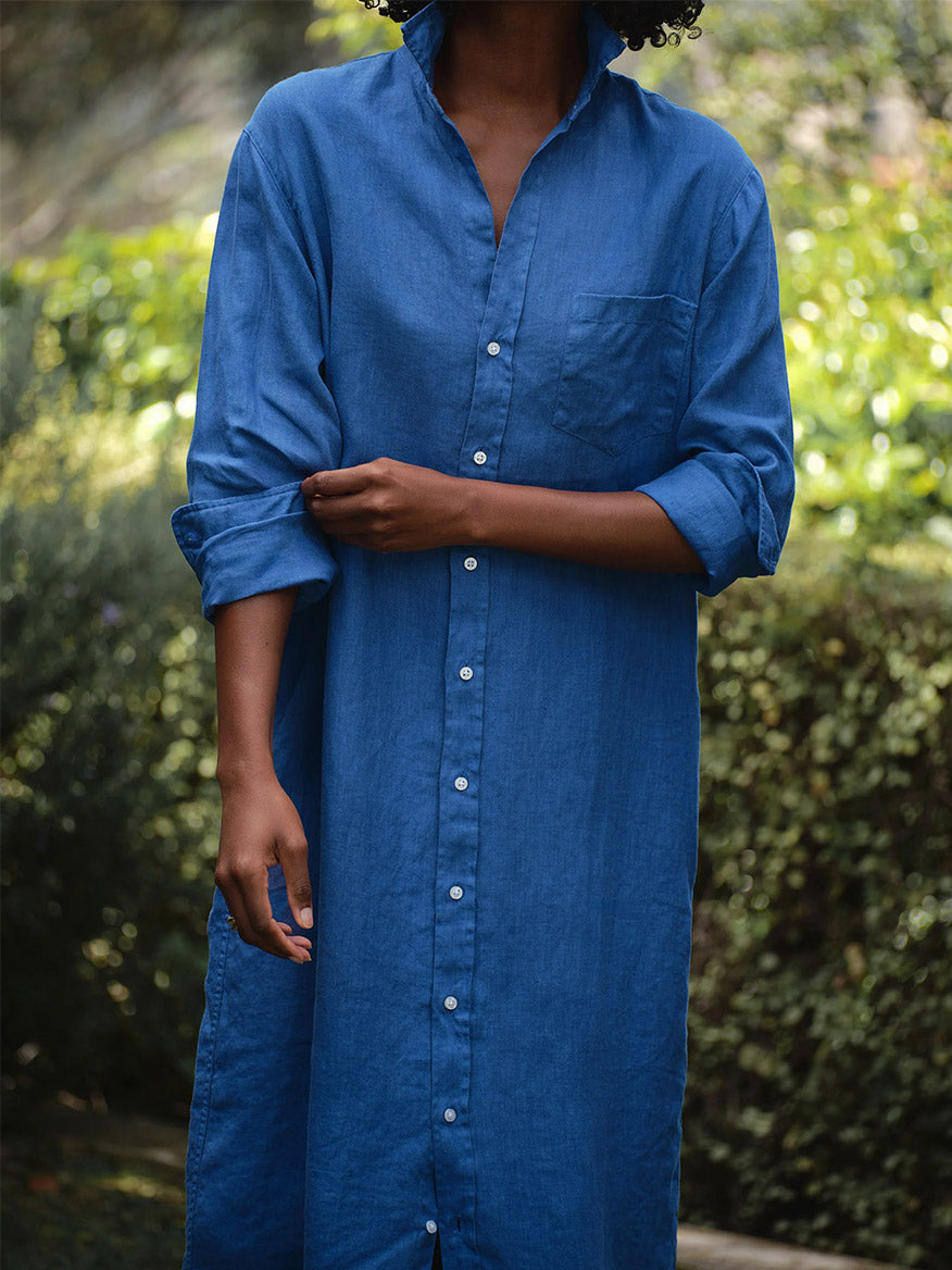 A person stands outdoors with greenery blurred in the background, effortlessly rolling up one sleeve of the Frank & Eileen Rory Maxi Shirtdress in Cabo Blue—a timeless touch of California chic in this classic button-down dress.