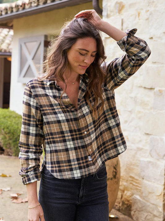 A woman in a Frank & Eileen Silvio Untuckable Button-Up Shirt in Sand & Camel with Black Plaid adjusts her hair while standing outside a building.