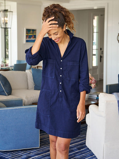 A woman stands in a living room, wearing a Frank & Eileen Southern Ireland Utility Mini Dress in Navy with six pockets and smiling with one hand on her head. She is in front of a sofa set and a door is visible in the background.