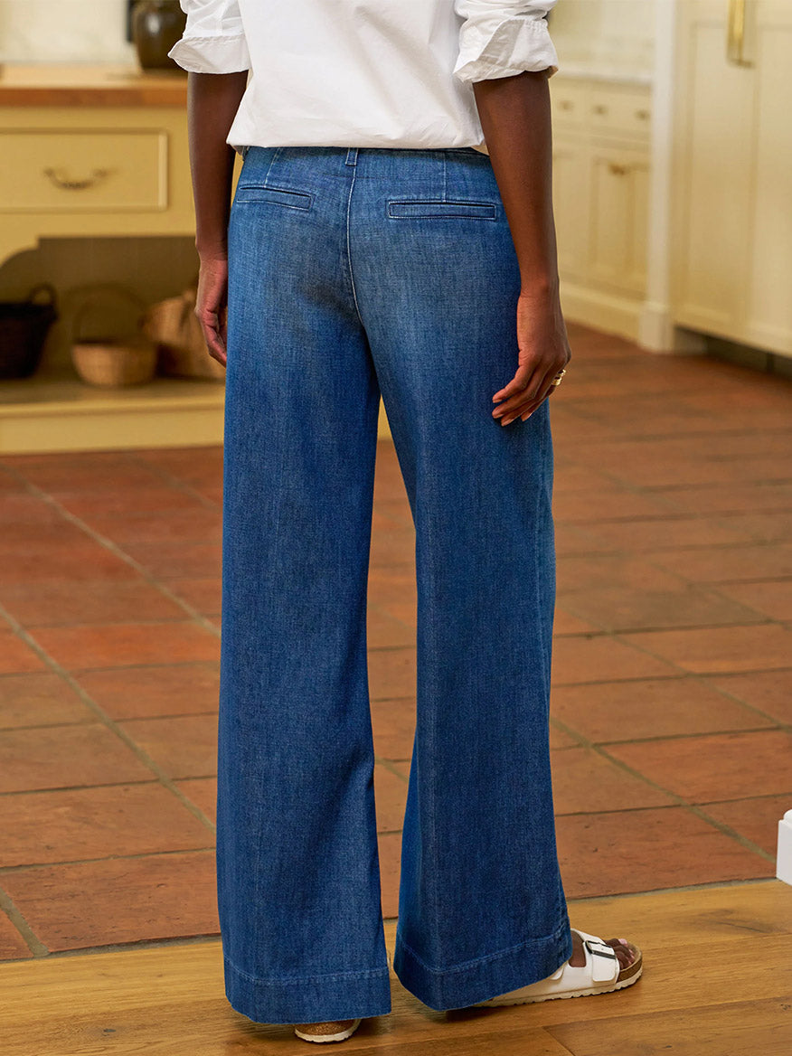 A person wearing Frank & Eileen West Village NYC Wide-Leg Trouser in 1984 Wash, a white top, and white sandals stands indoors on a tile and wood floor, facing away from the camera.