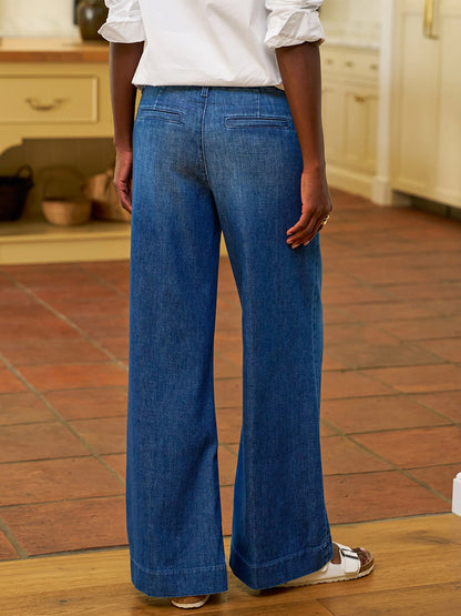 A person wearing Frank & Eileen West Village NYC Wide-Leg Trouser in 1984 Wash, a white top, and white sandals stands indoors on a tile and wood floor, facing away from the camera.