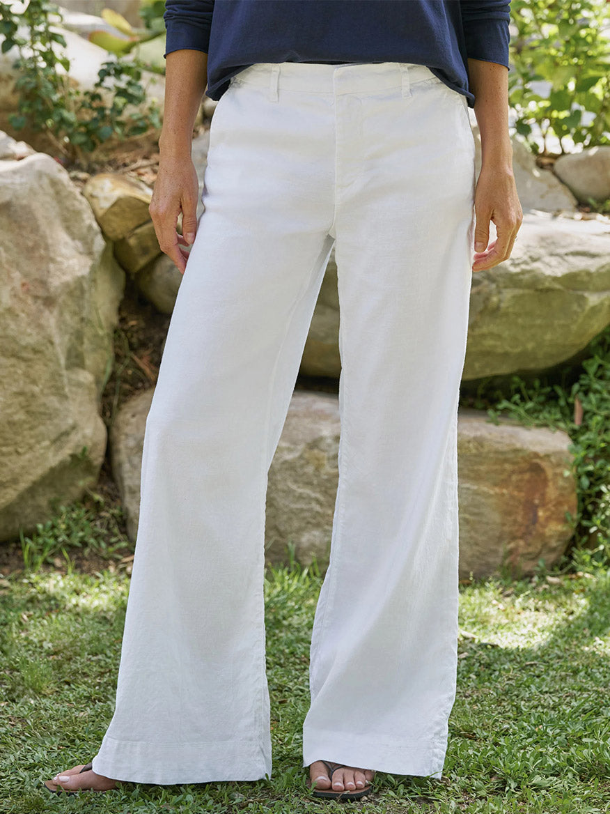 A person stands outdoors on grass, wearing Frank & Eileen West Village NYC Wide-Leg Trouser in White with a dark long-sleeve top and sandals. Only the lower body is visible, with large rocks and greenery offering a chic NYC-inspired backdrop.