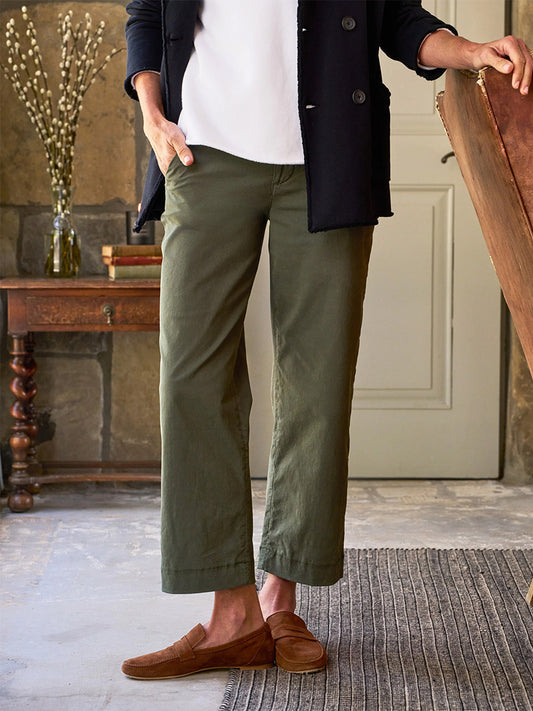 A person wearing the Frank & Eileen Westport Italian Wide-Leg Chino in Army, complemented by a black jacket and brown loafers, stands in a room adorned with a striped rug and antique wooden furniture.