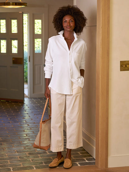 A person stands in the doorway, elegantly dressed in a white shirt and Frank & Eileen Westport Italian Wide-Leg Chino in Khaki featuring a contour waistband, holding a tote bag. The floor is tiled, and through the open door, lush greenery is visible outside.