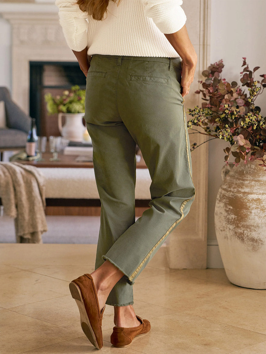 Wearing Frank & Eileen Wicklow Italian Chino in Army with Gold Tuxedo Stripe, a white sweater, and brown loafers, a person stands indoors near a large vase with flowers.