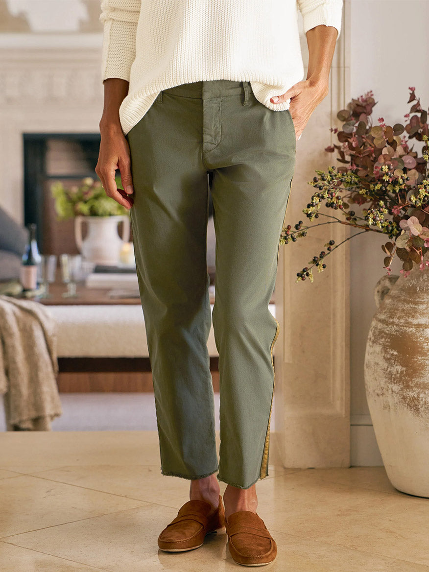 Indoors, a person wears the Frank & Eileen Wicklow Italian Chino in Army with Gold Tuxedo Stripe, paired with a white sweater and brown loafers, one hand in their pocket. A vase with branches appears in the background.