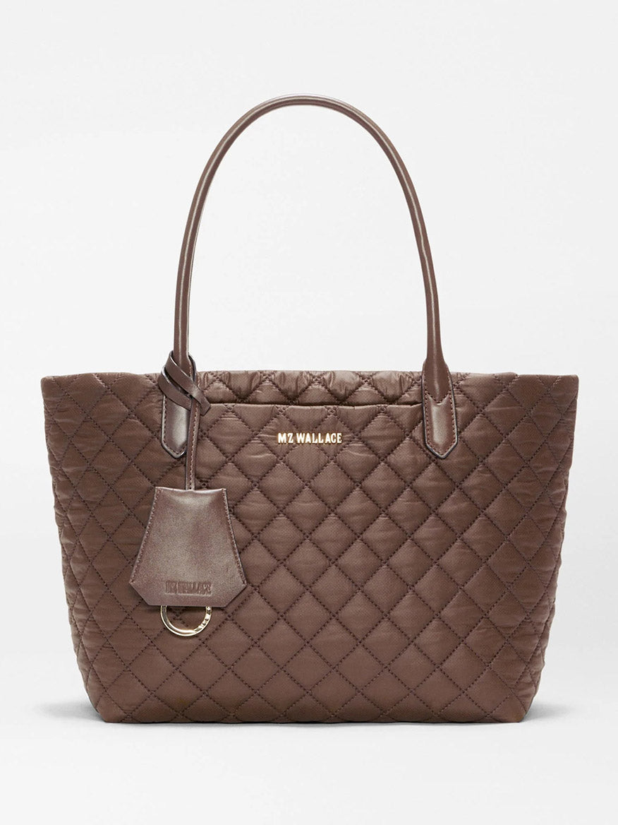 The MZ Wallace Small Daily Tote in Walnut Oxford is a brown quilted tote bag made from recycled nylon, featuring two Italian leather handles, gold hardware, and a logo tag. “MZ WALLACE” is printed in gold lettering near the top center of the bag.