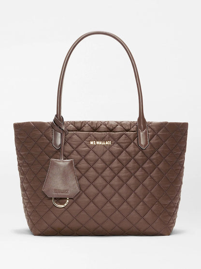 The MZ Wallace Small Daily Tote in Walnut Oxford is a brown quilted tote bag made from recycled nylon, featuring two Italian leather handles, gold hardware, and a logo tag. “MZ WALLACE” is printed in gold lettering near the top center of the bag.