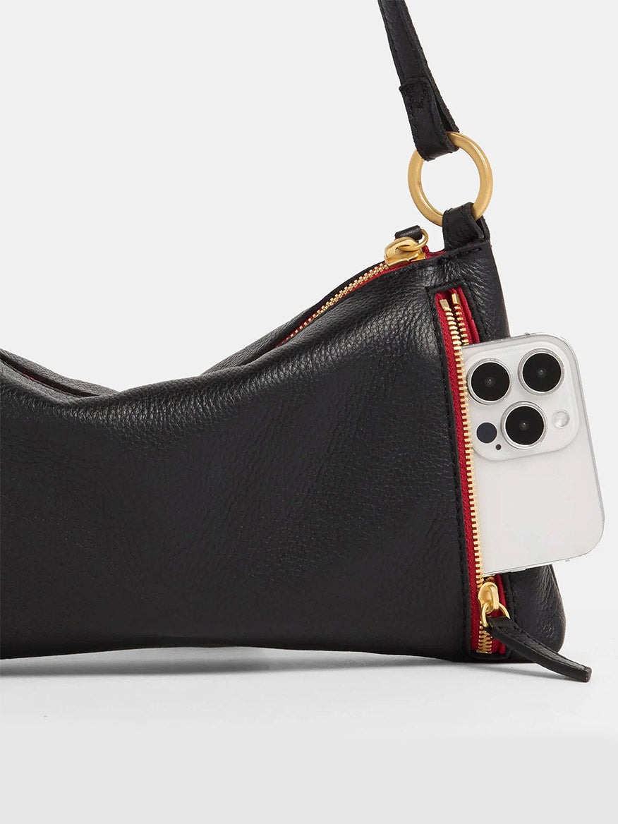 The Hammitt Los Angeles Kyle Shoulder Bag in Black/Brushed Gold & Red Zip is a versatile accessory crafted from black pebbled leather. It features a gold ring detail, a red zipper, and an adjustable crossbody strap. Its outer pocket conveniently holds a white smartphone.