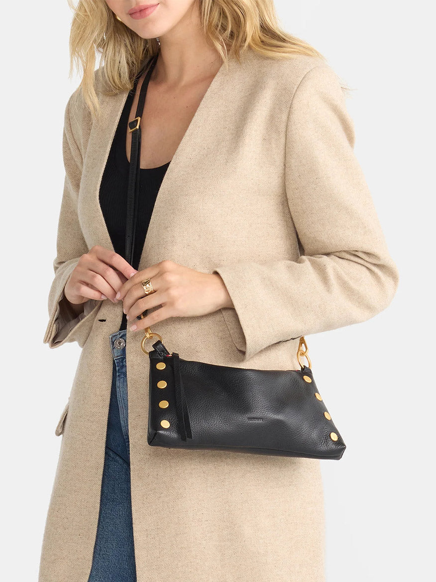 A person in a beige coat holding the Hammitt Los Angeles Kyle Shoulder Bag, crafted from black pebbled leather and featuring brushed gold embellishments with a red zip.