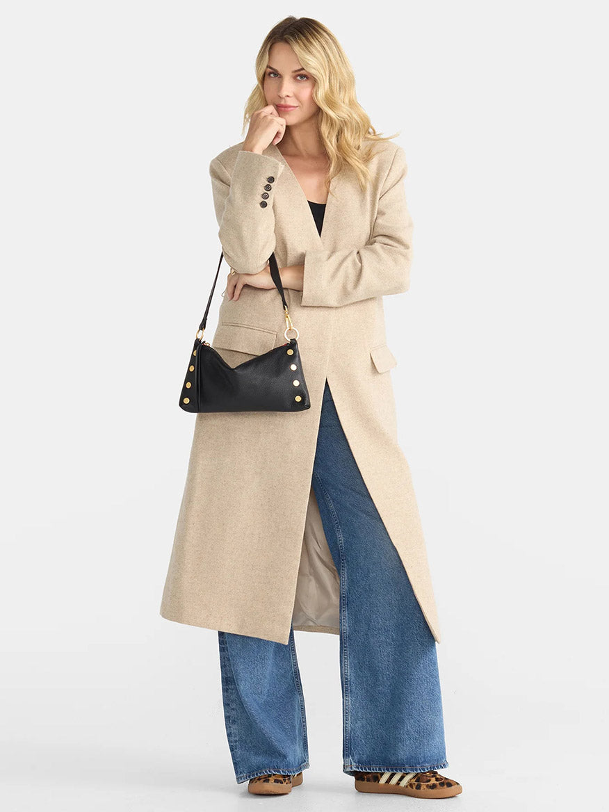 A person dressed in a long beige coat, blue jeans, and brown shoes is standing against a light background, carrying the Hammitt Los Angeles Kyle Shoulder Bag in black with brushed gold accents and a red zip, featuring an adjustable crossbody strap.