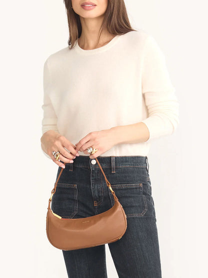 A woman wearing a cream sweater and dark jeans holds the Hammitt Los Angeles Becker Small crossbody bag in Mahogany Pebble, featuring brushed gold hardware.