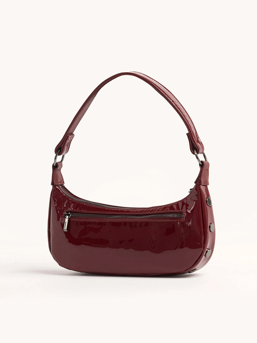 The Hammitt Los Angeles Becker Small in Pirouette Patent is a handbag crafted from fine Italian materials in a burgundy red hue. It features a single shoulder strap along with a removable, adjustable crossbody strap. The design is completed with a front zipper pocket and side button details for added style.