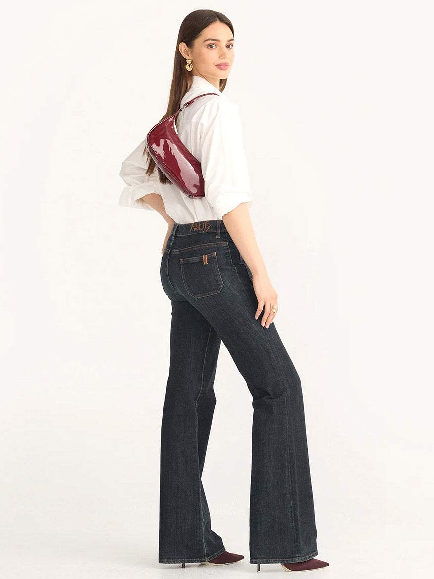 A person in a white shirt and dark flared jeans stands sideways, holding the Hammitt Los Angeles Becker Small in Pirouette Patent, a red shoulder bag featuring an Italian patent leather finish and a removable adjustable crossbody strap.