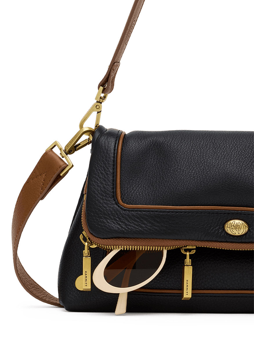 A black pebbled leather Hammitt Los Angeles Beverly Medium bag in East End/Antique Gold, detailed with brown trim and gold hardware. The design features a pair of sunglasses partially tucked into its external zippered pocket, and includes a convenient crossbody strap for hands-free versatility.