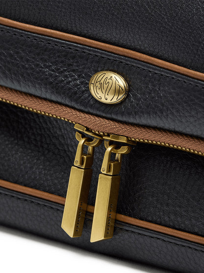 Close-up of the Hammitt Los Angeles Beverly Medium in East End/Antique Gold, featuring black pebbled leather with brown trim, two antique gold zippers, and an oval gold emblem with engraved detailing. Includes a crossbody strap for versatile carrying.