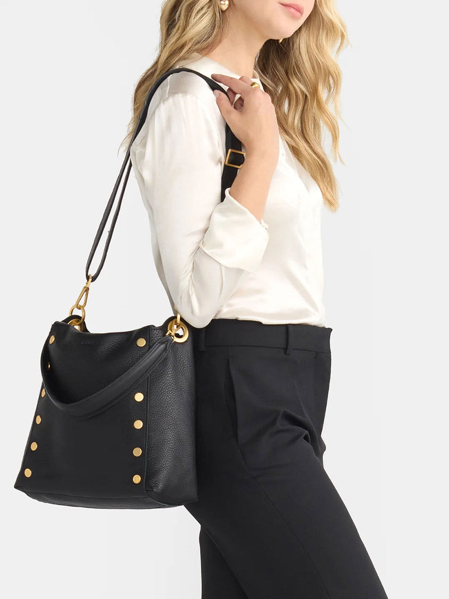 A person in a white blouse and black pants carries a Hammitt Los Angeles Bryant Large Crossbody Bag in Black & Brushed Gold, featuring gold studs.