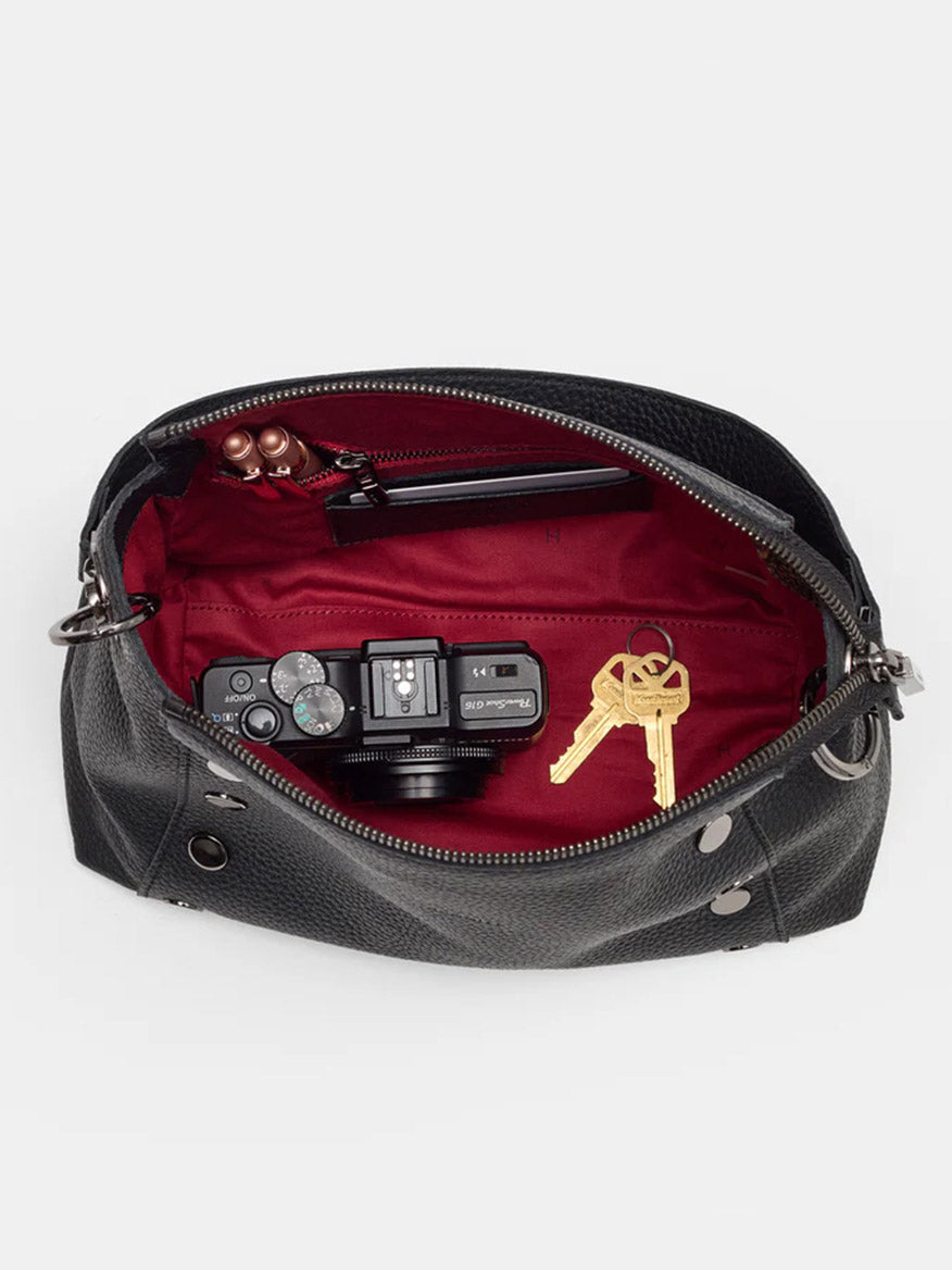 An open Hammitt Los Angeles Bryant Medium Crossbody Bag in black pebbled leather with a red interior reveals a camera, keys, and a pair of glasses.