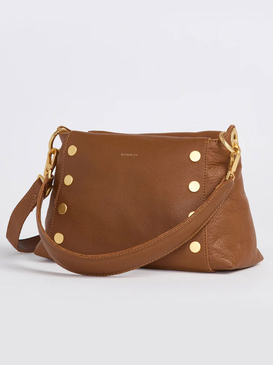 The Hammitt Los Angeles Bryant Medium Crossbody Bag in Mahogany Pebble boasts a luxurious brown leather exterior with a gold metal clasp and decorative gold studs. It features a wide strap connected by gold rings, along with an adjustable shoulder strap that adds to its versatile and elegant design, making it perfect for various occasions.