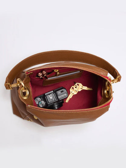 The Hammitt Los Angeles Bryant Medium Crossbody Bag in Mahogany Pebble features a brown design with a soft pebbled texture and a red interior that contains a camera, car keys, a few coins, and a small zippered pocket. The revamped Bryant Medium design also includes adjustable shoulder straps for added convenience.