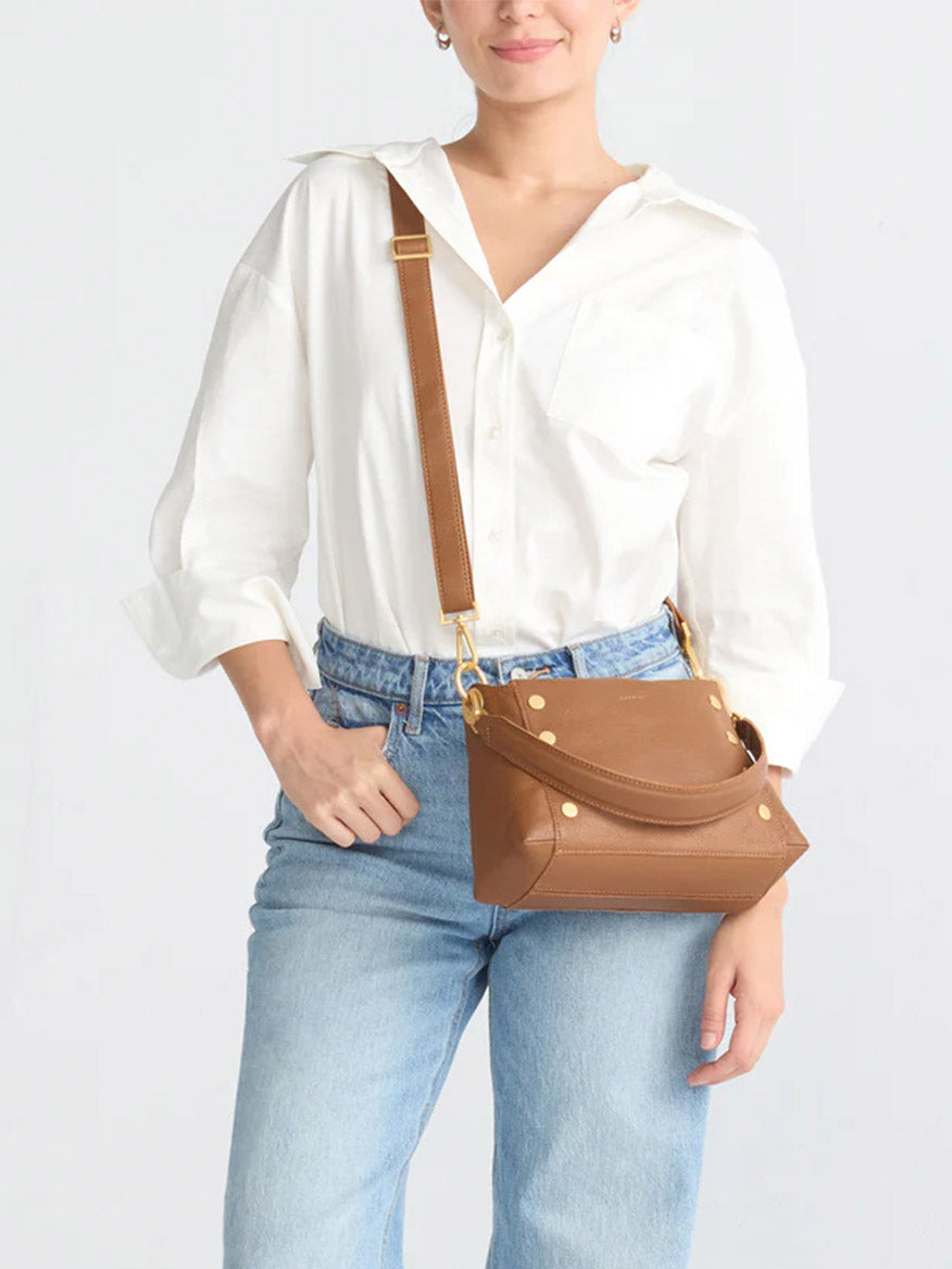 Person in a white shirt and blue jeans carrying the Hammitt Los Angeles Bryant Medium Crossbody Bag in Mahogany Pebble, featuring gold accents and adjustable shoulder straps.