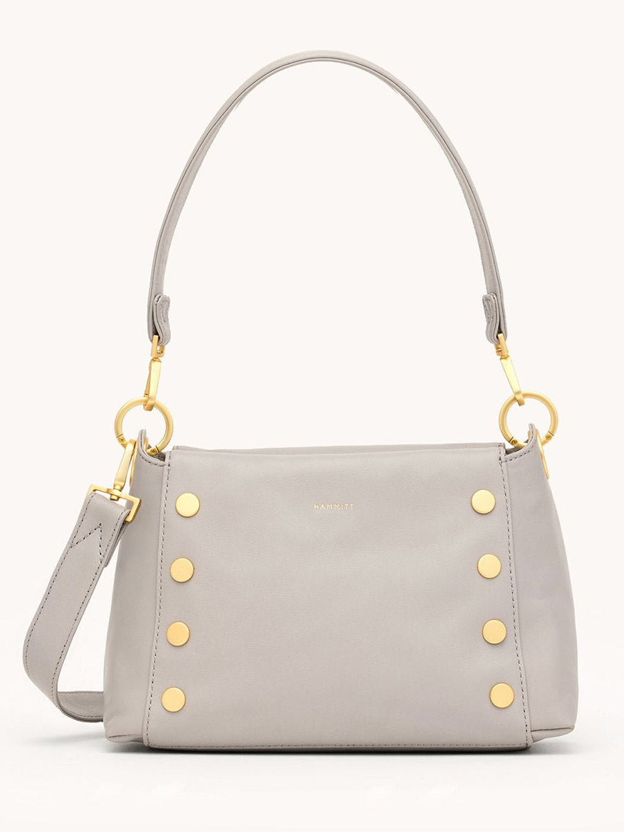 The Hammitt Los Angeles Bryant Medium Crossbody Bag in Stormy/Brushed Gold features light gray pebbled leather, gold studs, a detachable strap, and a short handle, shown against a plain background.
