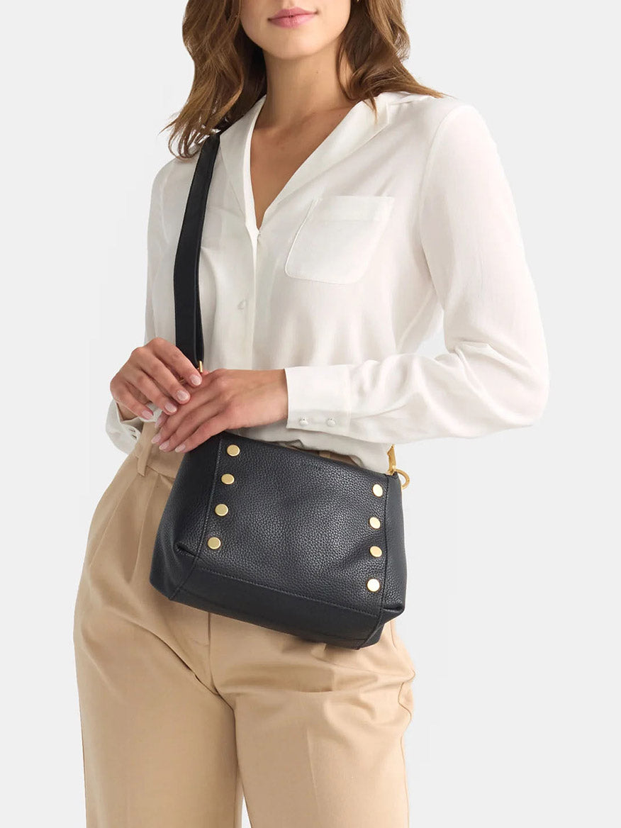 A woman in a white blouse and beige pants is holding the Hammitt Los Angeles Bryant Medium Crossbody Bag in black, detailed with gold studs, making it a versatile everyday accessory.